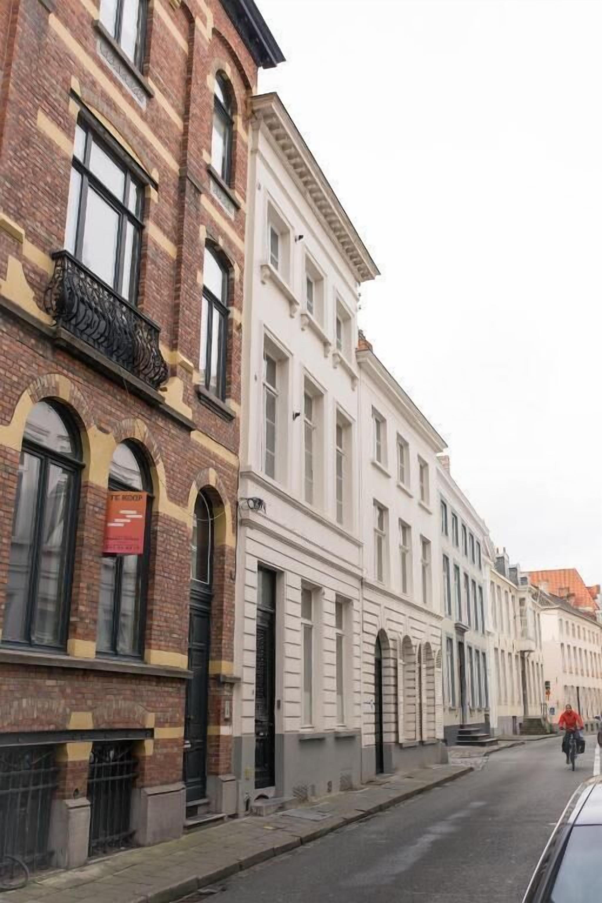 Calle con edificios históricos, incluyendo un edificio de ladrillo con balcón de hierro forjado negro y un edificio blanco con ventanales arqueados.