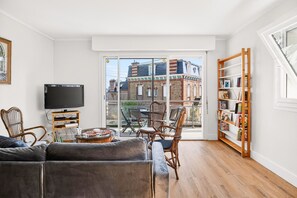 A living room with a television, a sofa, a dining table with chairs, a bookshelf, and a large window with a view of buildings. - Stunning sea and beach view for 6 (Dinard)