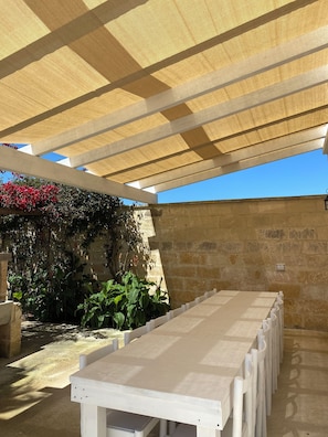 A covered outdoor area with a long white table, stone walls, and a pergola providing shade.