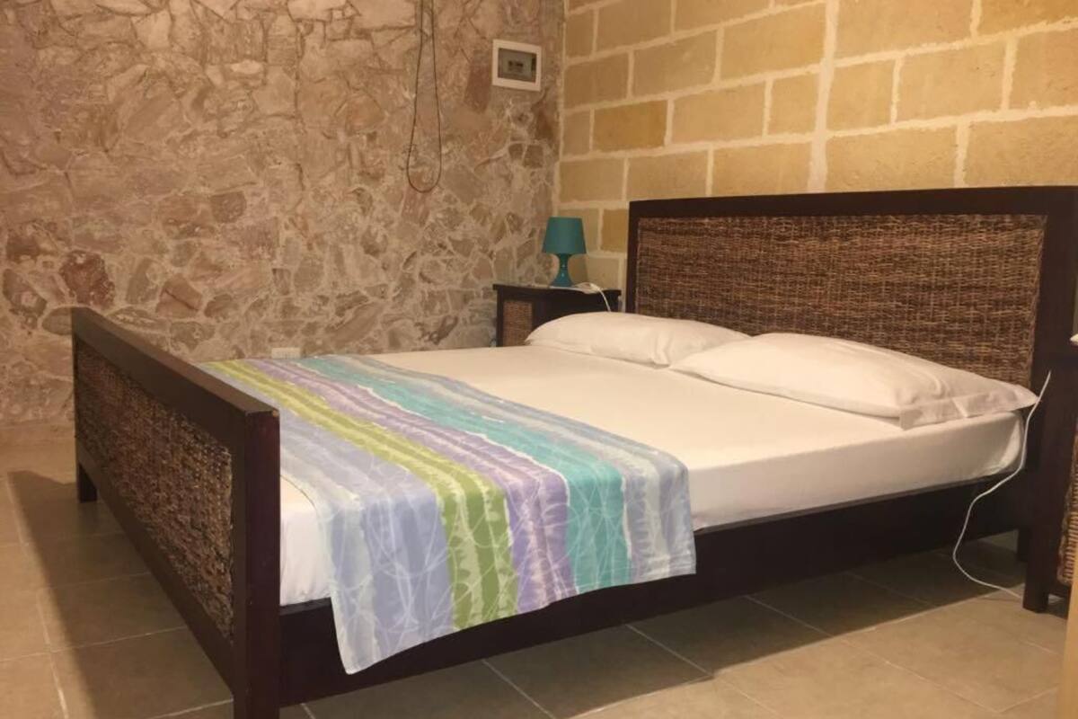 Un lit avec une couvre-lit à rayures, un mur en pierre, un mur en briques, une table de chevet avec une lampe et une prise de courant.
