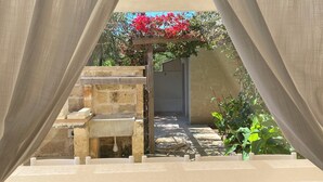A view through a window with sheer curtains, revealing a stone bench, a small garden with a pergola, and a wall with a water spout.