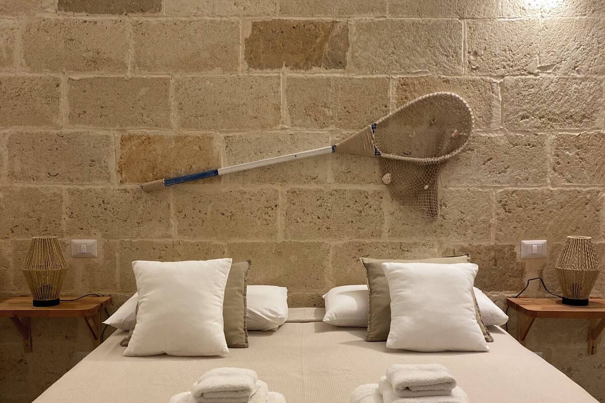 Un lit bien fait, avec du linge de lit et des serviettes blanches, une raquette de badminton accrochée au mur, et deux tables de chevet ornées d’objets décoratifs.