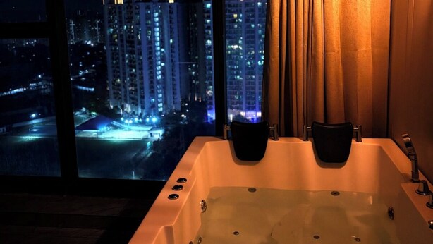 A modern bathroom with a large bathtub, a view of a cityscape at night, and a curtain. - Namaste B&B With Jacuzzi I Noida (Greater Noida)
