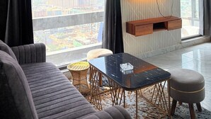 A modern living room with a sofa, a coffee table, a TV mounted on the wall, and large windows with curtains. - Namaste B&B With Jacuzzi I Noida (Greater Noida)