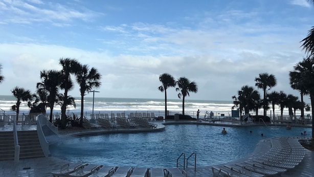 Ein Poolbereich mit Liegestühlen, Palmen und Blick aufs Meer.