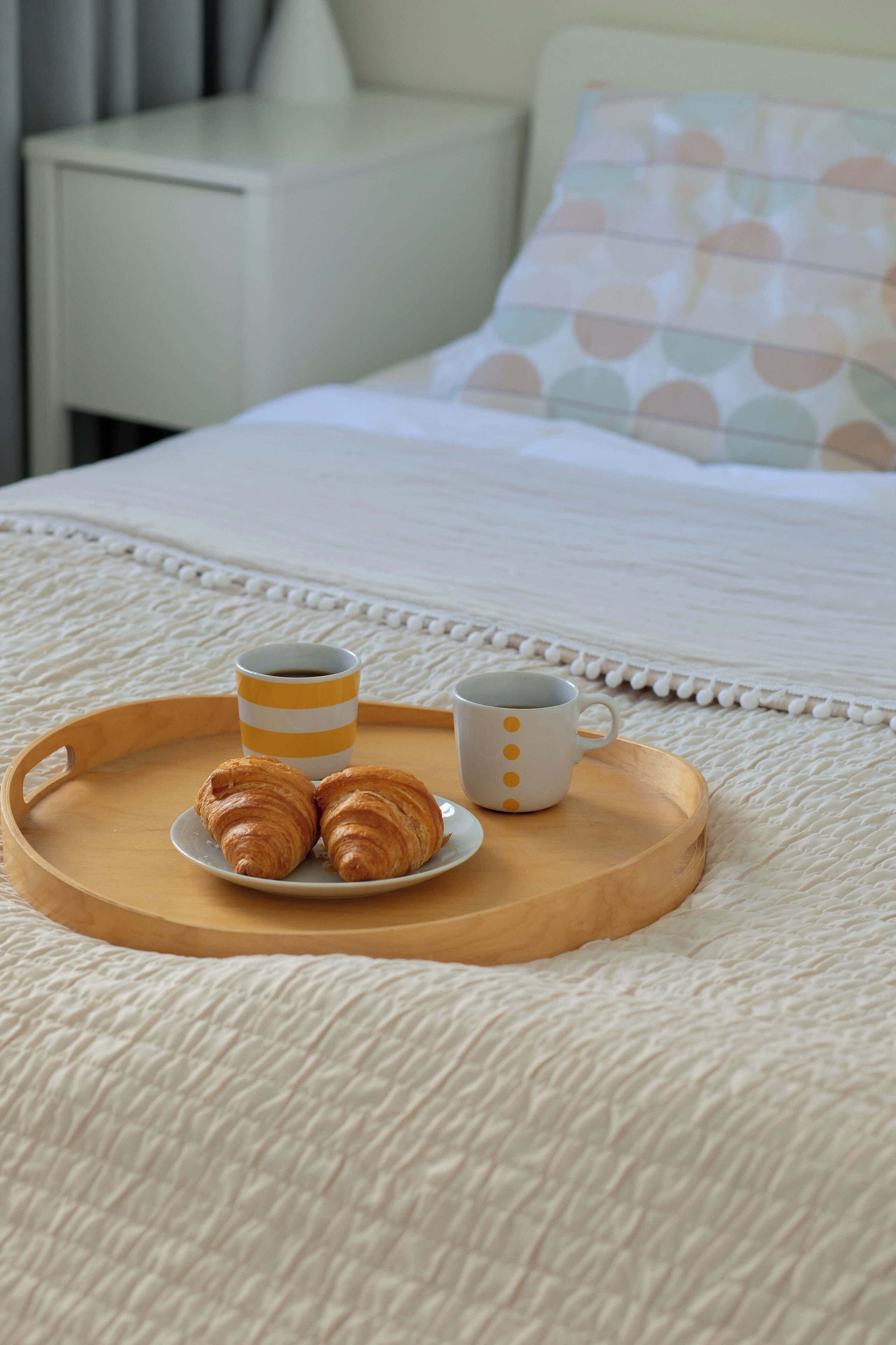 Ein Bett mit weißer Decke, ein Holztischchen mit Croissants und zwei Tassen, und ein weißer Nachttisch im Hintergrund.
