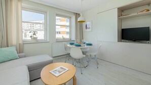 En moderne stue med sofa, spisebord dækket til to personer, fjernsyn og et vindue med gardiner.