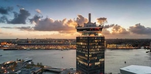 A high-rise building with a distinctive top, overlooking a body of water with boats and a cityscape in the background.
