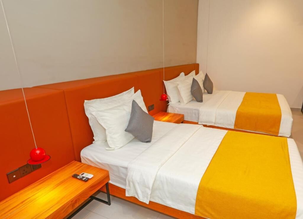 Una habitación de hotel con dos camas, cada una con ropa de cama blanca y un detalle amarillo. Hay una mesita de noche de madera con una lámpara roja y un control remoto.