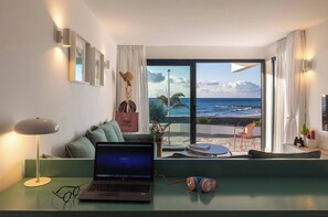 Ein modernes Zimmer mit einem Laptop auf einem Schreibtisch, Blick auf den Ozean durch große Fenster und einem grünen Sofa.