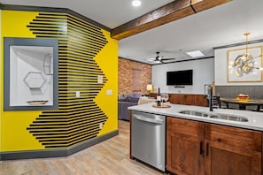 A modern kitchen with a yellow accent wall, a dishwasher, wooden cabinets, and a dining area with a table and chairs. - Carolina Condo - Pets Welcome - Walkable Downtown (Wilmington)