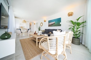 A modern living room with a sofa, dining table, wicker chairs, a potted plant, and a painting of a boat on the wall. - 1.8 Amapola Blue Sea: recently refurbished, in front of the (Maspalomas)