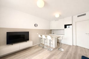 A modern kitchen with white cabinets, a flat-screen TV, and a bar area with white stools. - Wave Island 9 - Carraro Immobiliare Jesolo (Lido di Jesolo)