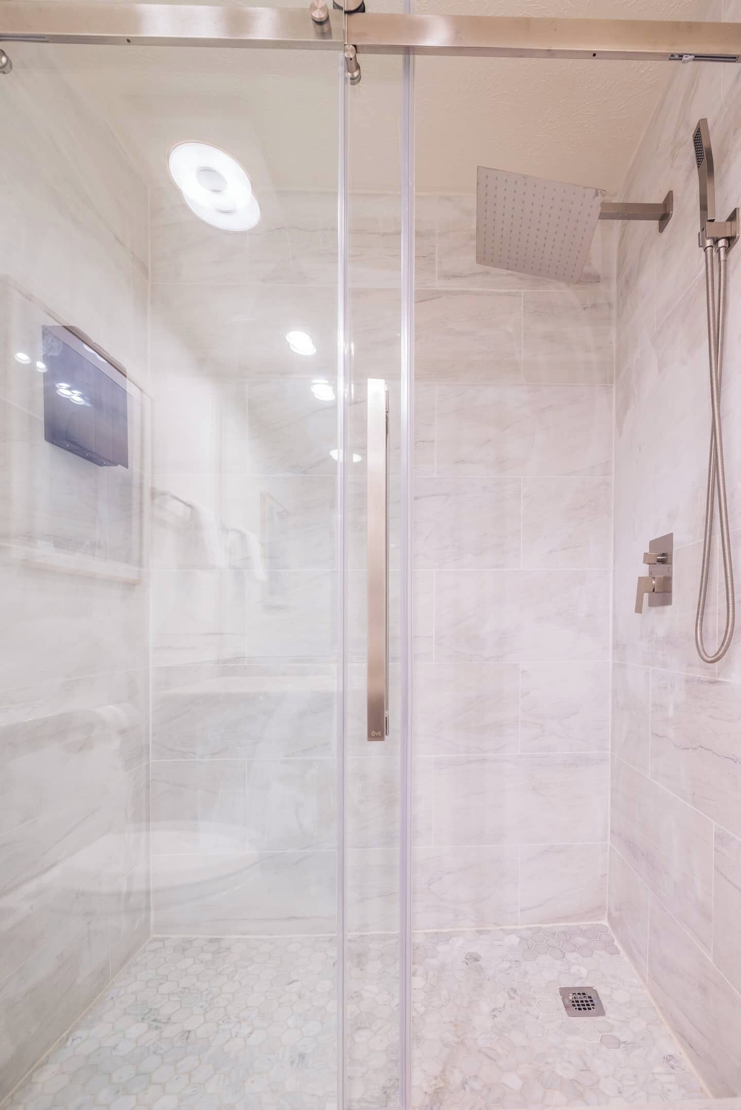 A glass-enclosed shower with a handheld showerhead, a wall-mounted control panel, and a tiled floor.