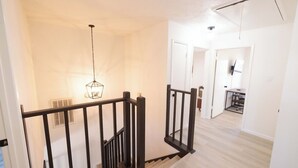 Una escalera con barandilla negra, una lámpara colgante y vista a una habitación con escritorio y silla.