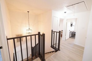Una escalera con barandilla negra, una lámpara colgante y vista a una habitación con escritorio y silla.