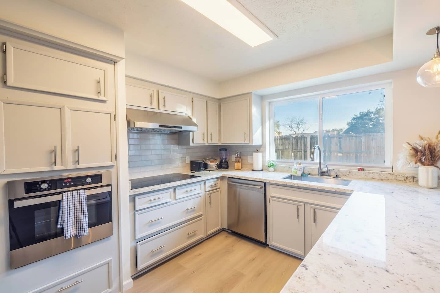 A modern kitchen with white cabinetry, a built-in oven, a dishwasher, and a large window providing natural light.