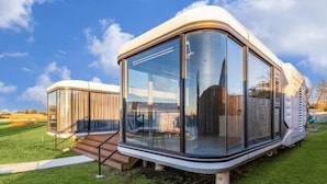 A modern RV with a large sliding glass door, a small porch area, and a grassy yard. - One-of-a-kind tiny home designed for an unforgettable stay by the beautiful San Jacinto River (Highlands)