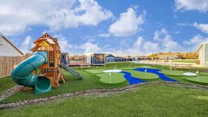 En lekplats med en rutschkana och en liten byggnad, en golfbana med flaggor och en klarblå himmel.