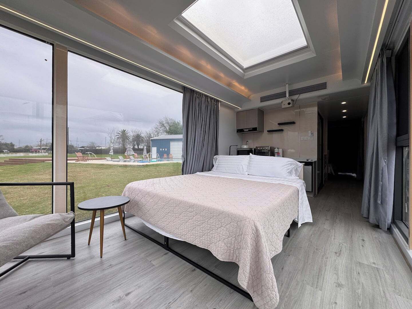 Quarto de hotel moderno com uma cama grande, uma mesinha, uma cadeira, cozinha compacta e vista para o exterior.