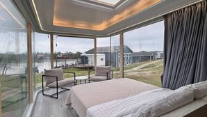 Ett sovrum med ett stort fönster, en säng, en sittgrupp och utsikt över en sjö och hus.