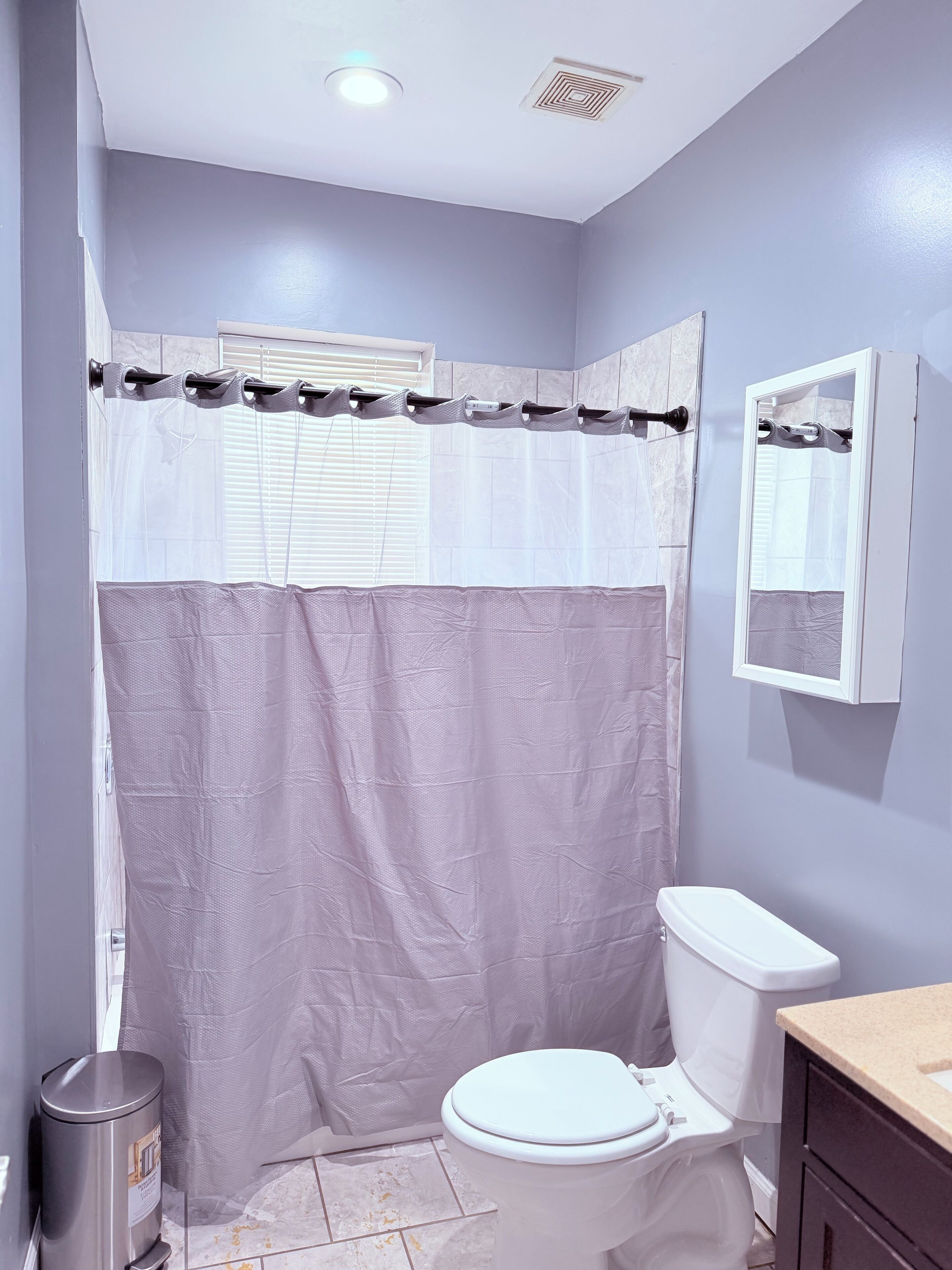 Ein Badezimmer mit Toilette, Mülleimer, Duschvorhang und einem Fenster mit Jalousien.