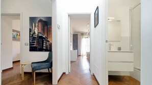 A hallway with a herringbone floor, a chair, a painting, and a bathroom with a mirror and sink. - Albertelli - Large and comfortable apartment for 6 (Roma)
