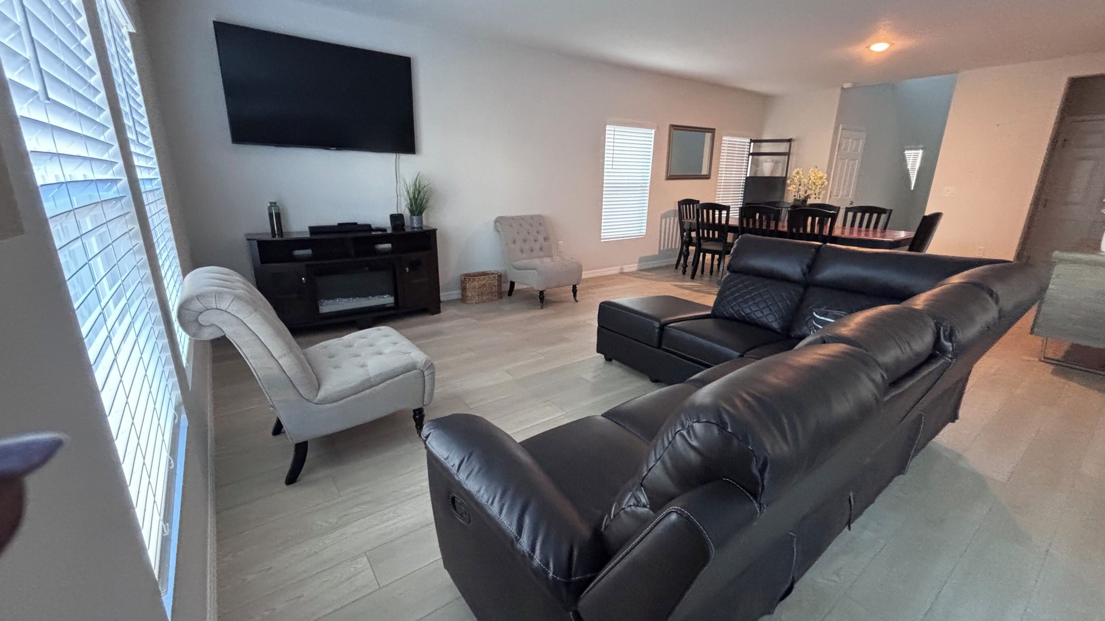 A modern living room with a black leather sofa, a white armchair, a flat-screen TV, a dining area with a table and chairs, and a large window with blinds.