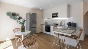 Een moderne keuken met witte kasten, een roestvrijstalen koelkast, een magnetron en een koffieapparaat. Er is een eethoek met rieten stoelen en een witte tafel.
