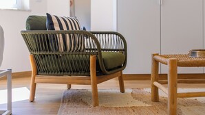 Een rieten stoel met kussens, een houten tafel en een geweven vloerkleed in een kamer met een witte muur en houten vloer.