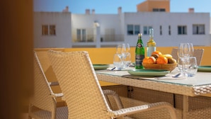 A dining table with chairs, a bottle, glasses, and a bowl of fruit on a balcony with a view of buildings. - Atlântico Azul - Apartment E (Lagos)
