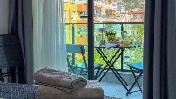 Quarto de hotel com varanda, uma mesa com frutas e um jarro, e uma toalha dobrada na cama.