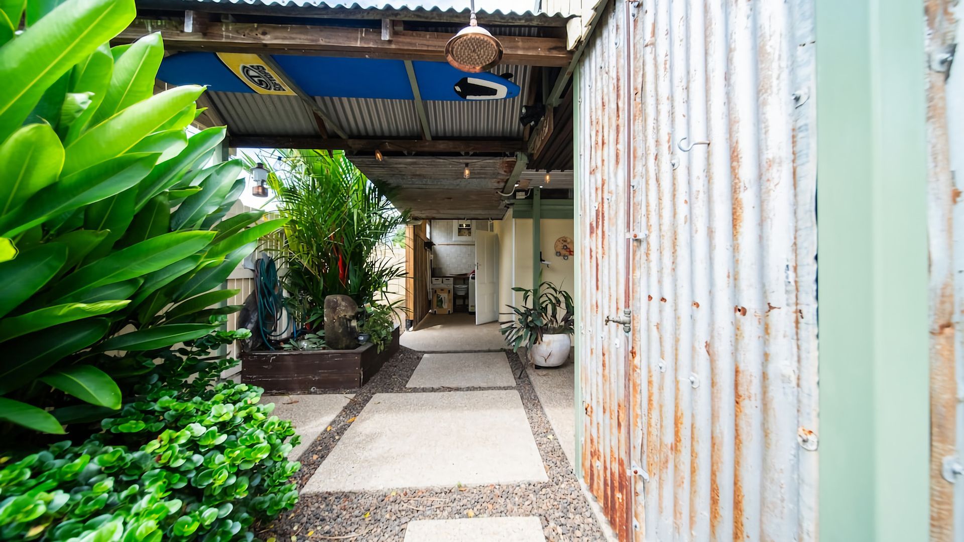 Un étroit sentier mène à un bâtiment rustique à l’extérieur recouvert de tôles ondulées, entouré d’une végétation luxuriante et d’un petit jardin.