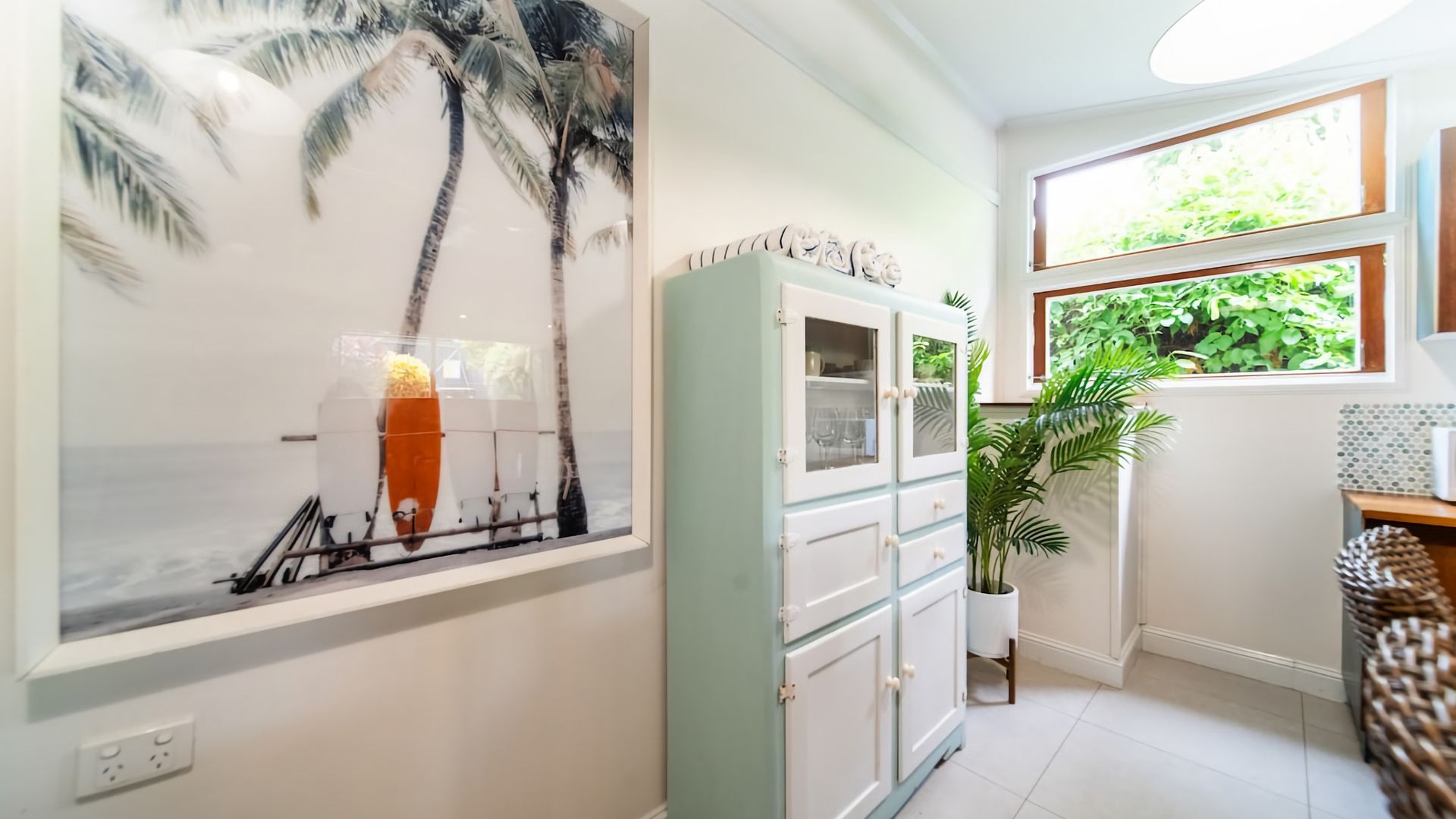 A room with a beach-themed wall art, a cabinet with glass doors, a potted plant, and a window with a view of greenery.