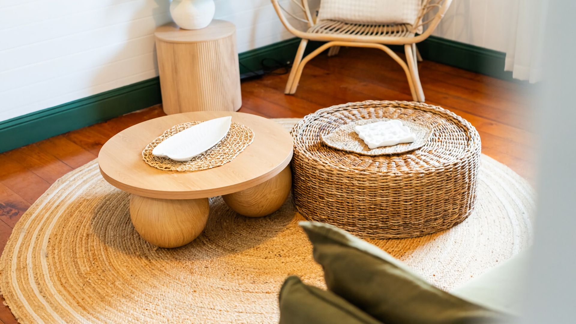 A round wooden table with a woven placemat and a woven ottoman with a cushion.