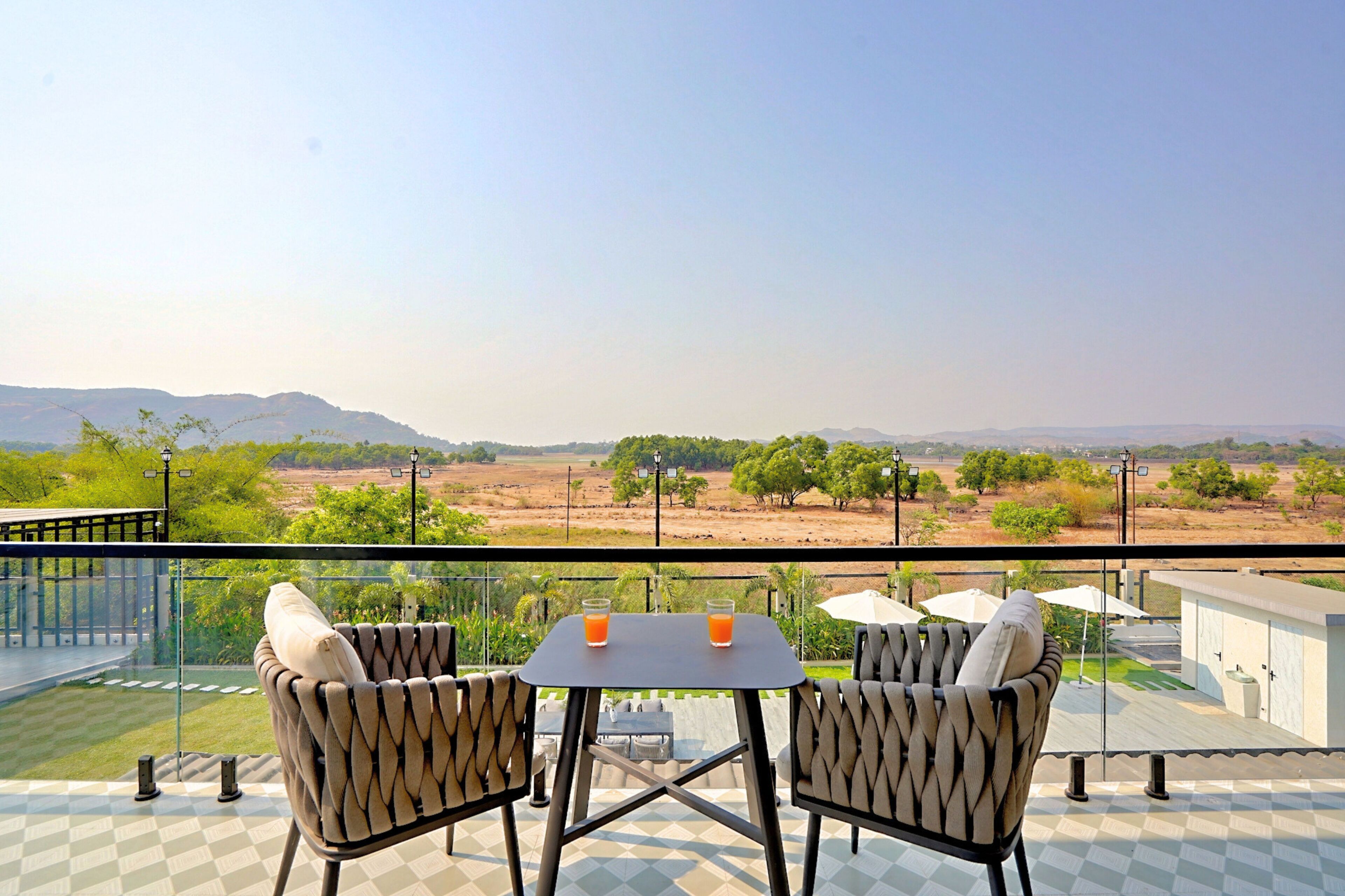 A balcony with two chairs and a table, two glasses of orange juice, and a view of a landscape with trees and mountains.
