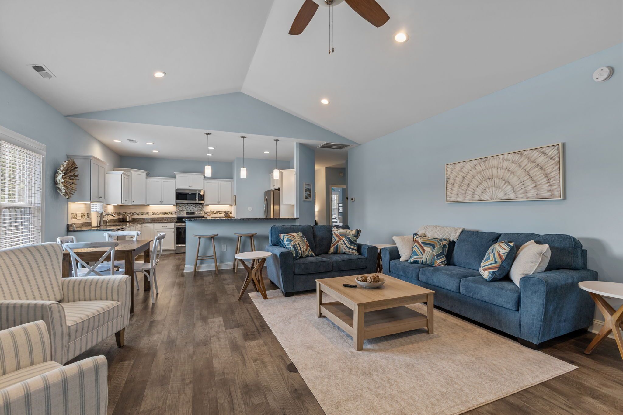 Un salón amplio con un sofá azul, sillones a rayas, una mesa de centro de madera y una cocina al fondo.