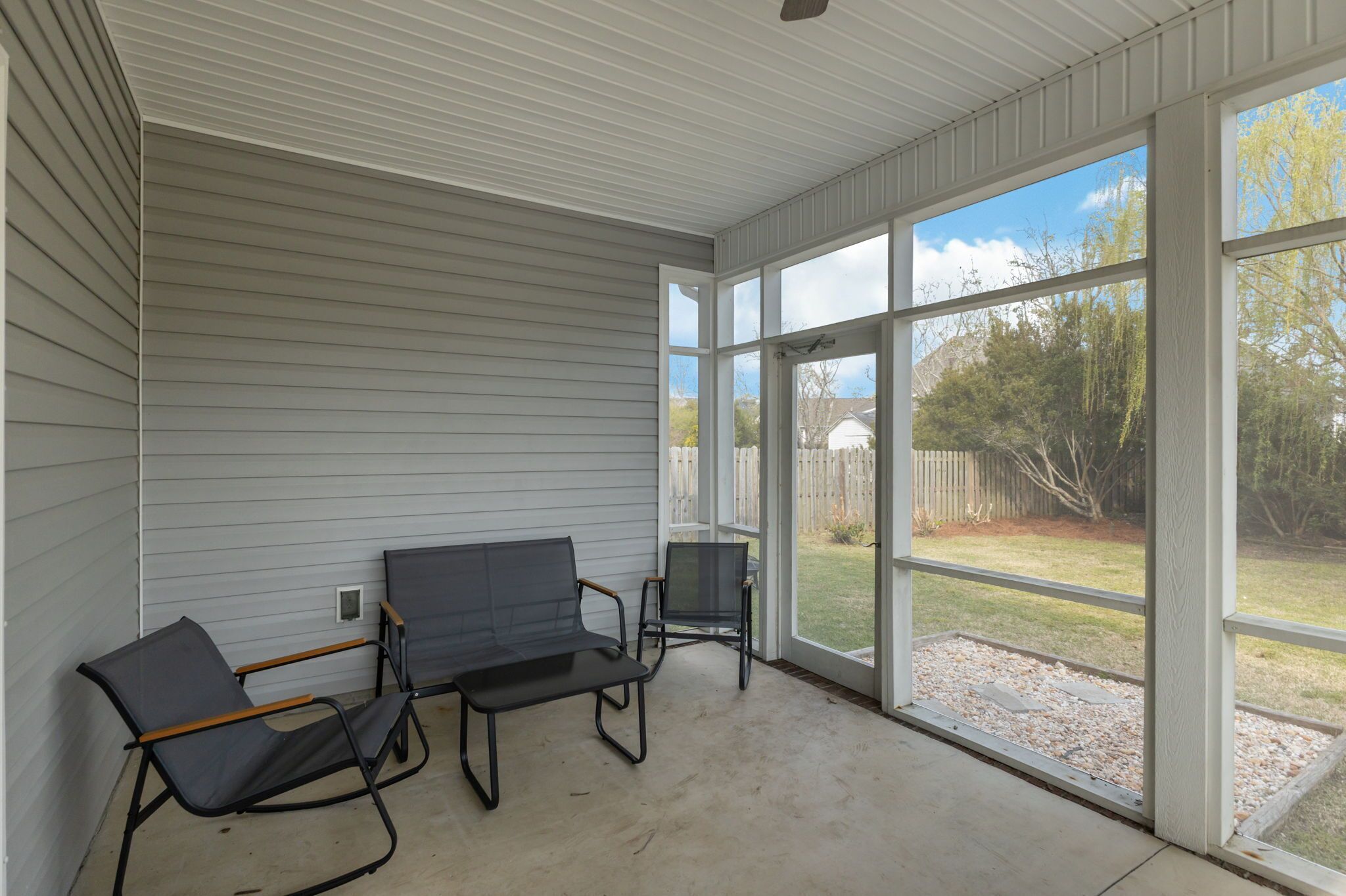 A covered porch with two chairs and a table, large windows, and a view of a backyard.