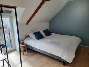 A bedroom with a sloped ceiling, a bed with white bedding and blue pillows, a small stool, and a window with a view. - Holiday Home 'Gite Les Copains - L'autre' with Private Terrace, Private Garden and Wi-Fi (Bourseul)