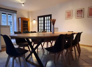 Un coin repas avec une table ronde, des chaises, un vase contenant des fleurs séchées, une bibliothèque et une fenêtre avec des stores.