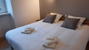 Een net opgemaakt bed met kussens en opgevouwen handdoeken, een nachtkastje met een lamp, en een raam met zonwering.