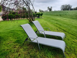 Deux fauteuils de couleur grise sont placés dans un espace extérieur verdoyant, agrémenté d’arbres, avec un bâtiment visible à l’arrière-plan.