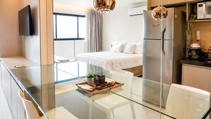 Une chambre d’hôtel moderne dotée d’une table à manger en verre, d’un lit avec du linge de maison blanc, d’un coin cuisine et d’un téléviseur à écran plat fixé au mur.