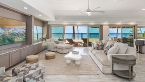 A spacious living room with a large flat-screen TV, comfortable seating, a ceiling fan, and a view of the ocean.