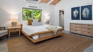 A bedroom with a large bed, two bedside lamps, a wooden headboard, a window with a view of greenery, a wooden dresser, and two wall-mounted artworks.