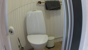 Un baño con inodoro, un cubo de basura pequeño, un perchero con toalla y un dispensador de papel higiénico.