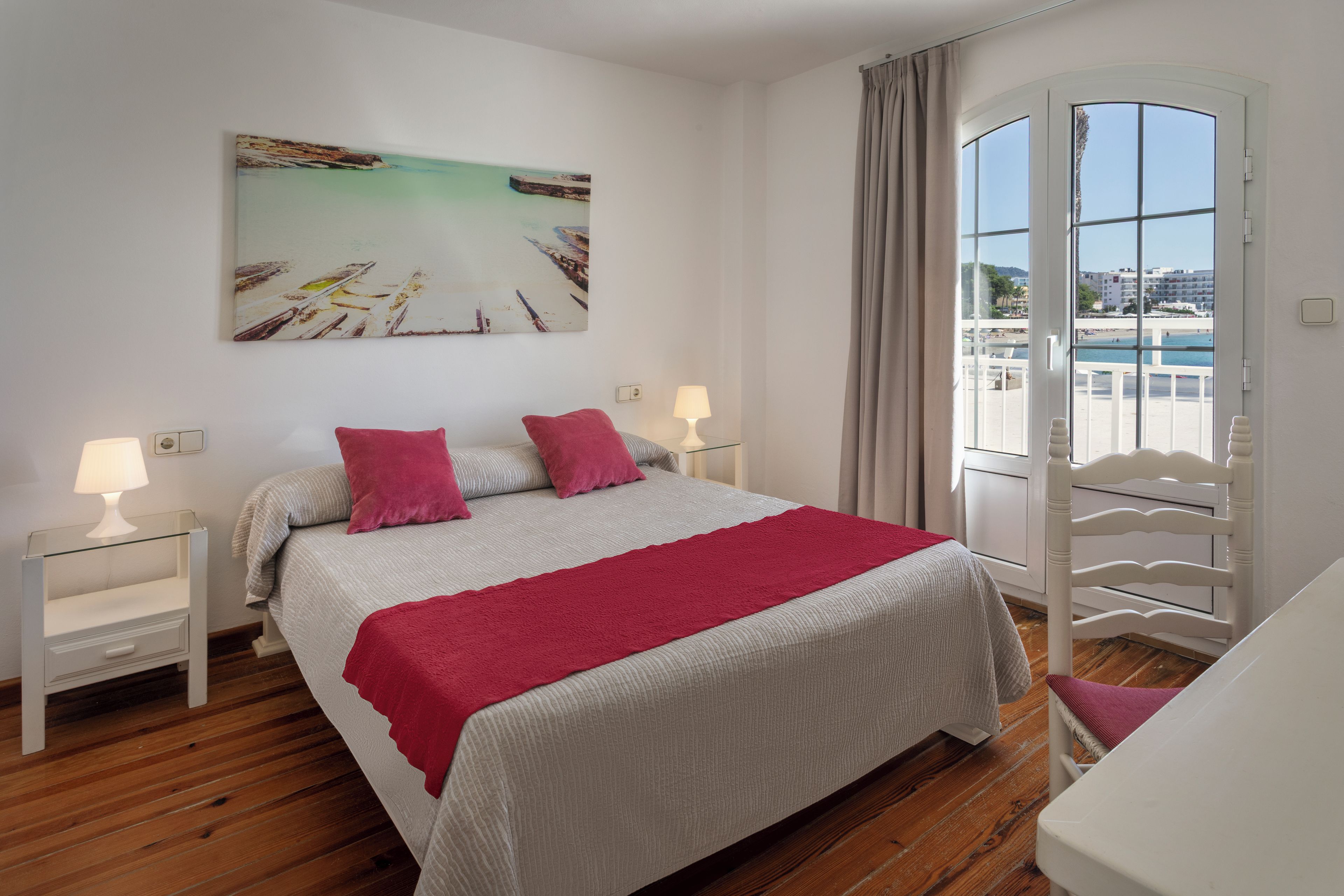 A bedroom with a bed, two bedside tables, a window with a view of a beach, and a painting on the wall.