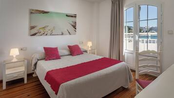 A bedroom with a bed, two bedside tables, a window with a view of a beach, and a painting on the wall.
