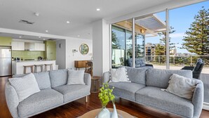 A modern living room with two grey sofas, a coffee table with vases, a kitchen in the background, and large windows offering a view of the outside. - Stylish 2-bed With Terrace, 3-min Walk to Beach (Adelaide)