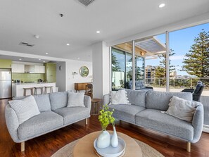 Ein modernes Wohnzimmer mit zwei grauen Sofas, einem Couchtisch mit Vasen, einer Küche im Hintergrund und großen Fenstern mit Blick nach draußen.